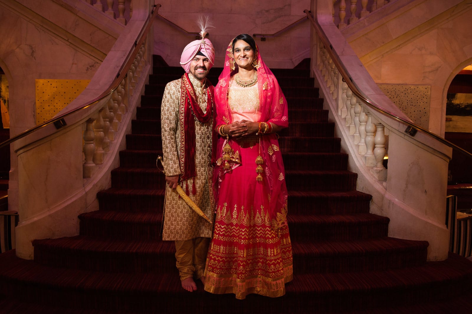 A groom wearing a gold kaftan and pink turban holds a sword while standing with his bride who is wearing a red and gold sari after their Renaissance Phipps South Asian Wedding.