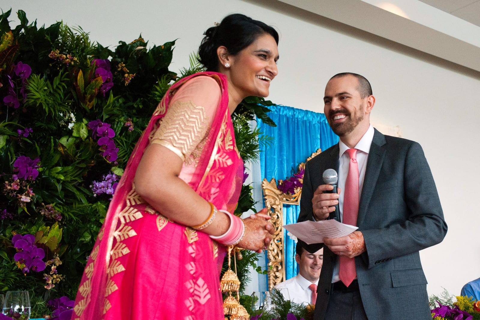 A bride in a chartreuse and gold sari laughs with her groom wearing a grey suit as they express greetings to their guests at their Renaissance Phipps South Asian Wedding.