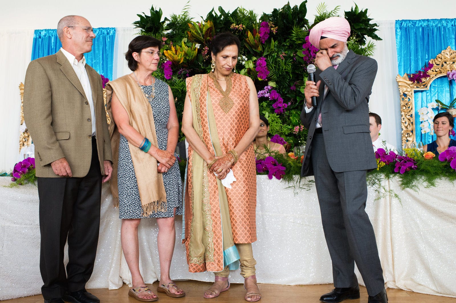A turban-wearing father of the bride wipes a tear from his eye as he stands with his wife and the groom's parents while offering a toast to the bride and groom during their Renaissance Phipps South Asian Wedding.