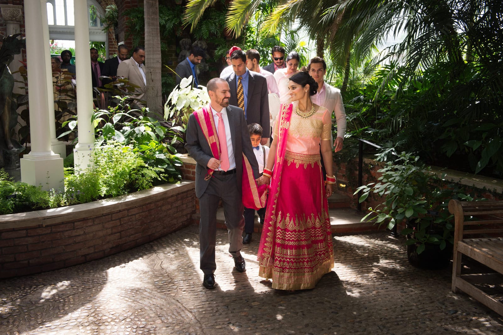 A groom wearing a grey suit and pink tie walks with his chartreuse and gold wearing bride as they lead their guests through a lush room in a conservatory during their Renaissance Phipps South Asian Wedding.