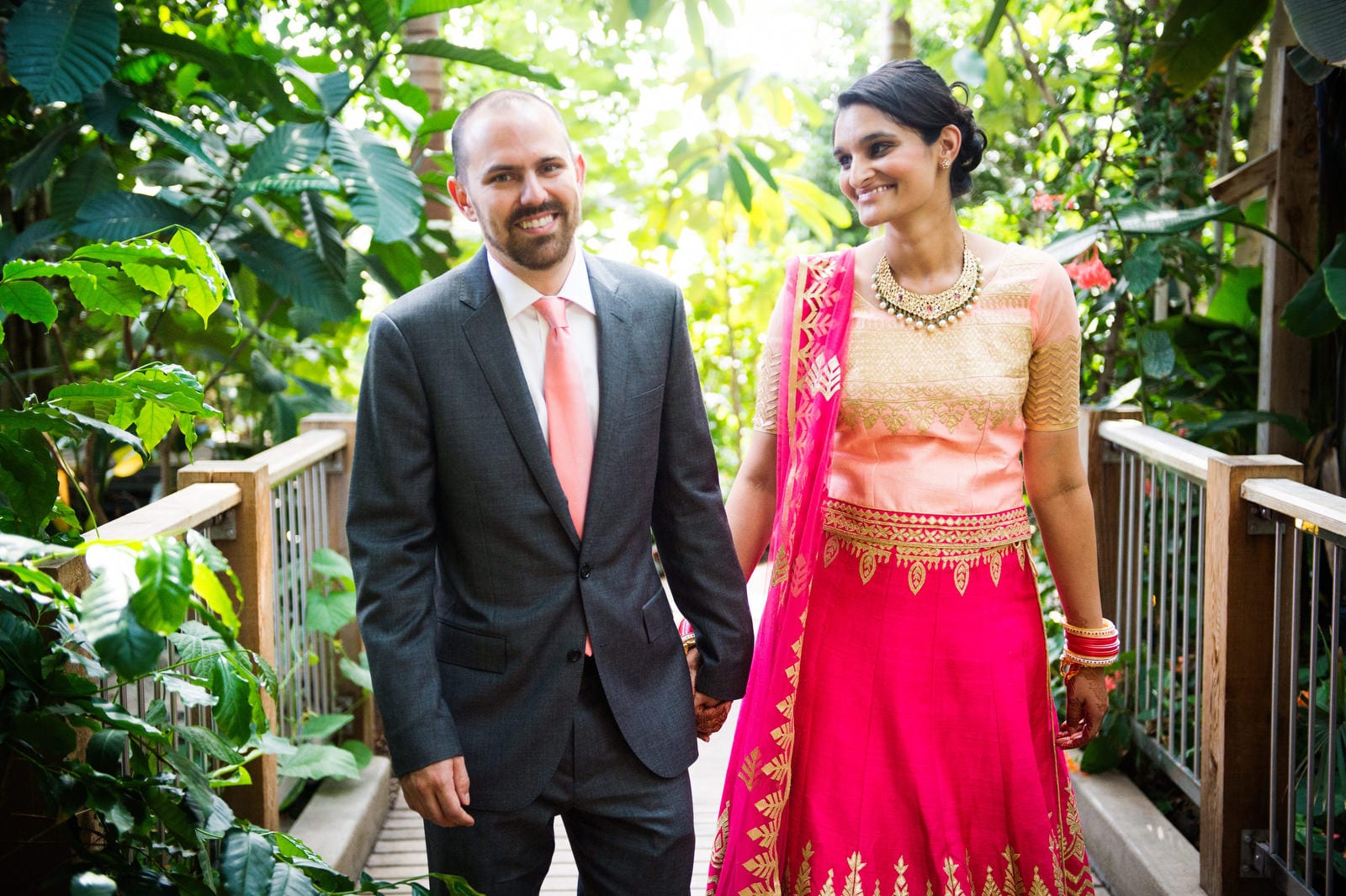 A bride and groom hold hands and smile as they stand in a greenhouse after their Renaissance Phipps South Asian Wedding.
