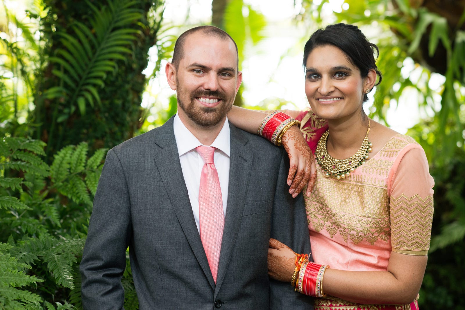 A bride in a gold sari rests her arm on her grooms shoulder. He's wearing a gray suit with a pink tie during their Renaissance Phipps South Asian Wedding.
