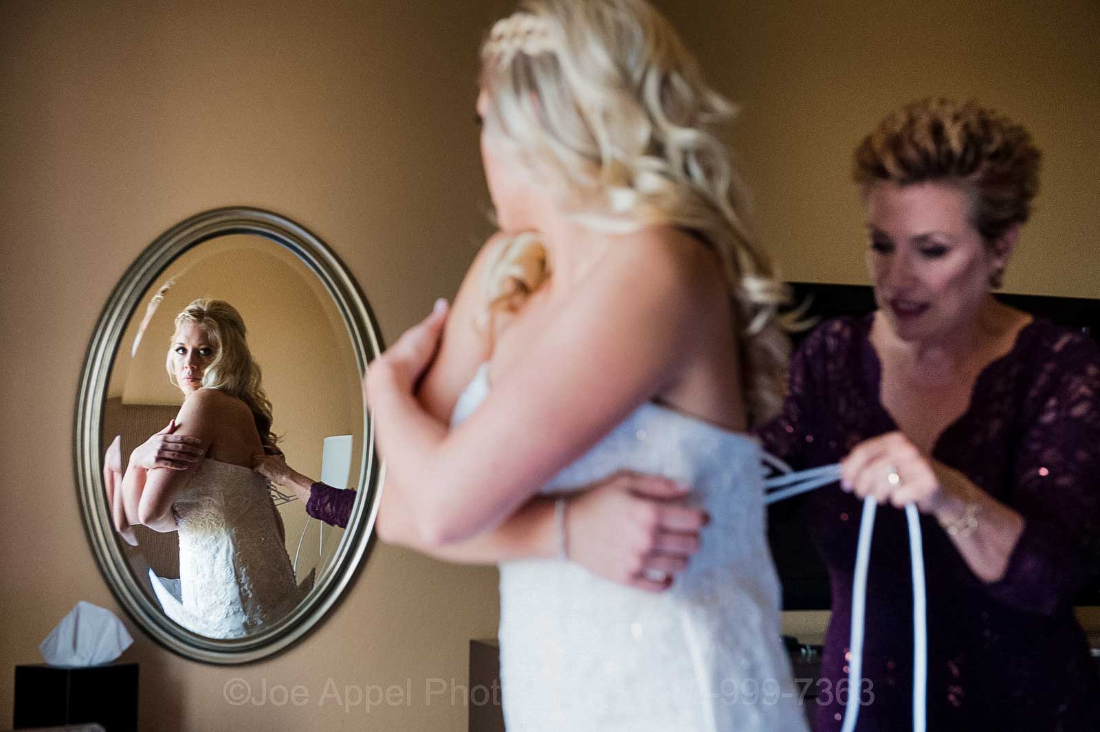 A blonde bride holds her shoulders and looks into an oval mirror as her mother ties the laces of her wedding dress at the Embassy Suites hotel near the Pittsburgh airport.