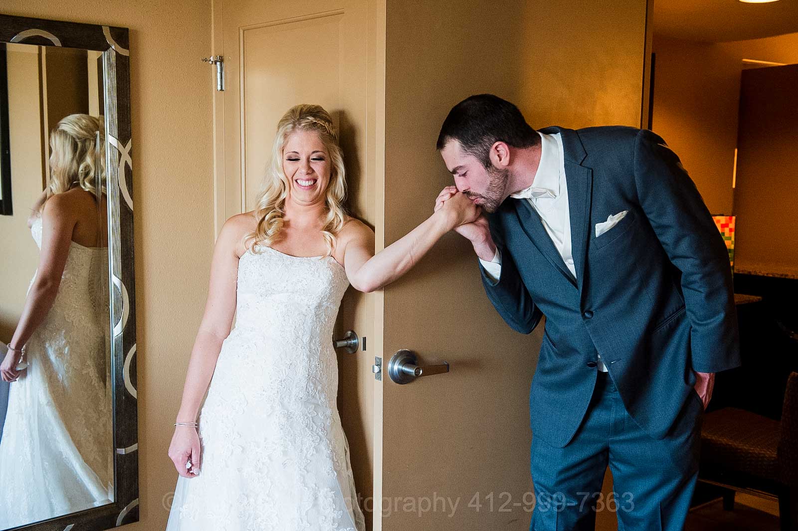 A bride smiles as her groom kisses her hand from around the corner of a door before their wedding at the Embassy Suites near Pittsburgh International Airport.