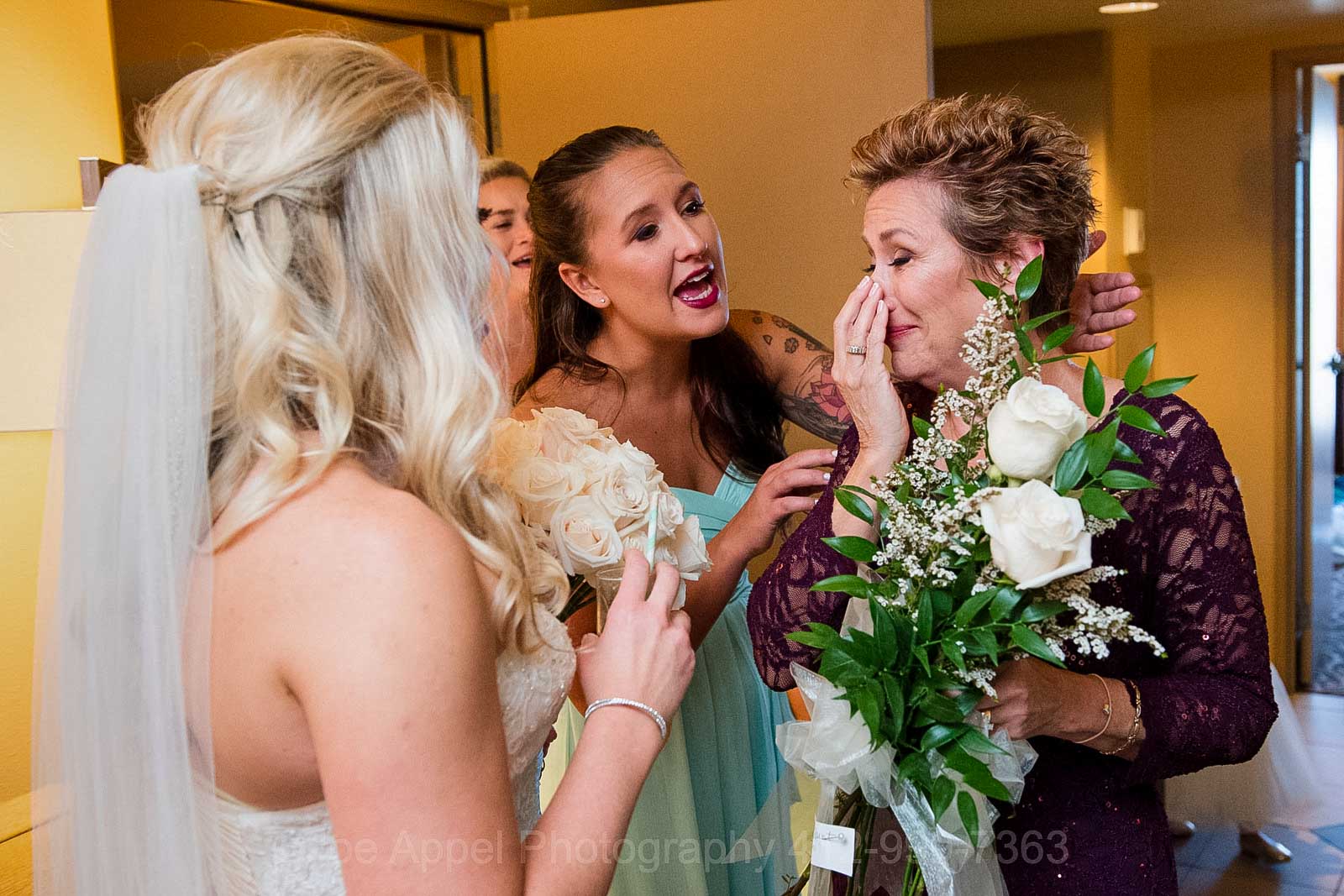 Bridesmaids and a bride comfort the bride's mother as she cries on seeing her daughter.