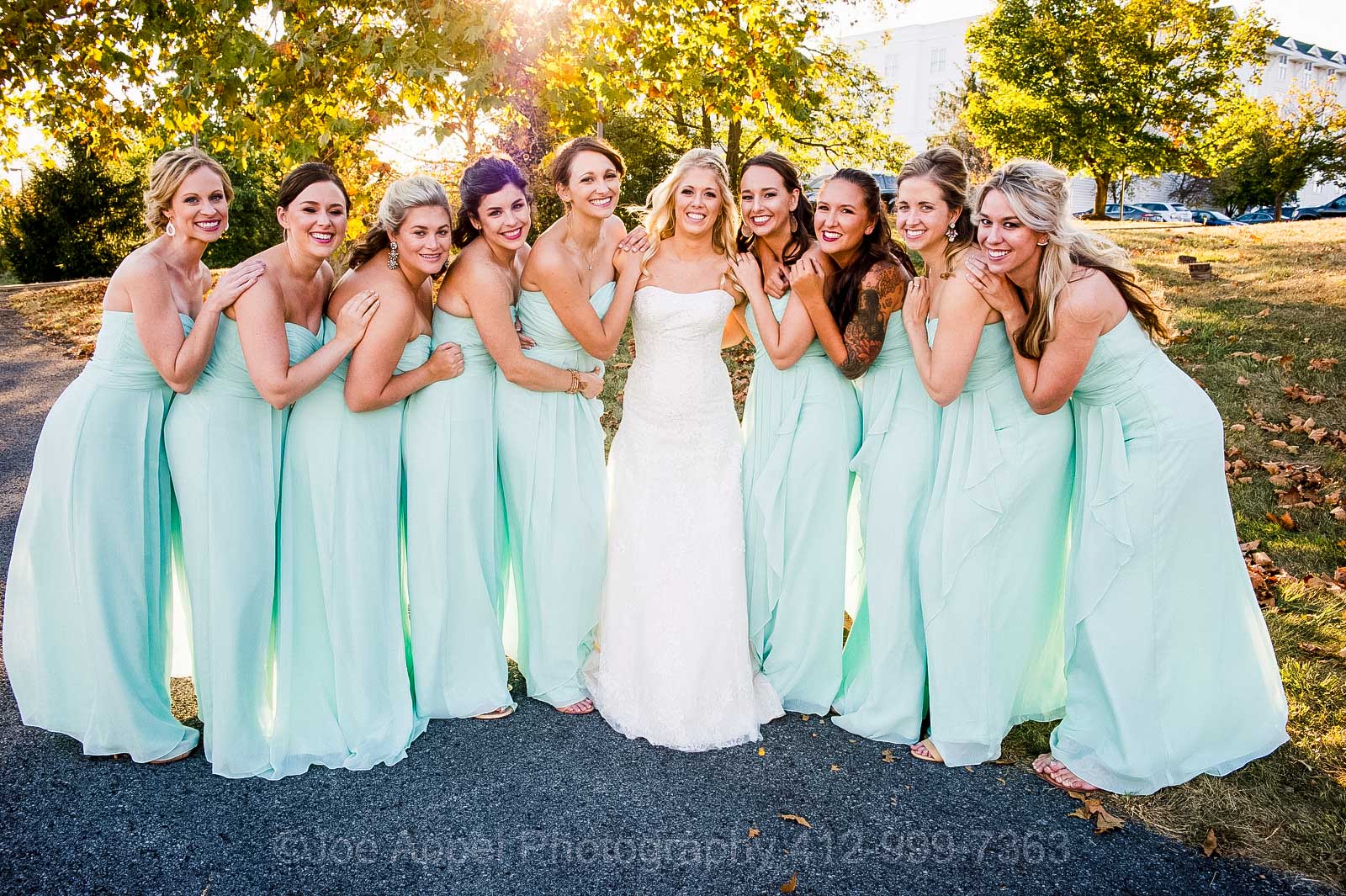 A bride is surrounded by bridesmaids in light blue-green dresses with the sunlight behind them.