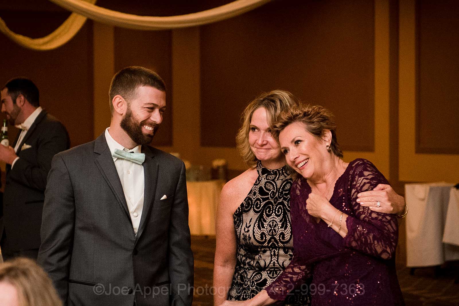 The bride's mom smiles as she is embraced by another woman while standing with her son as they watch the bride dance with her father.