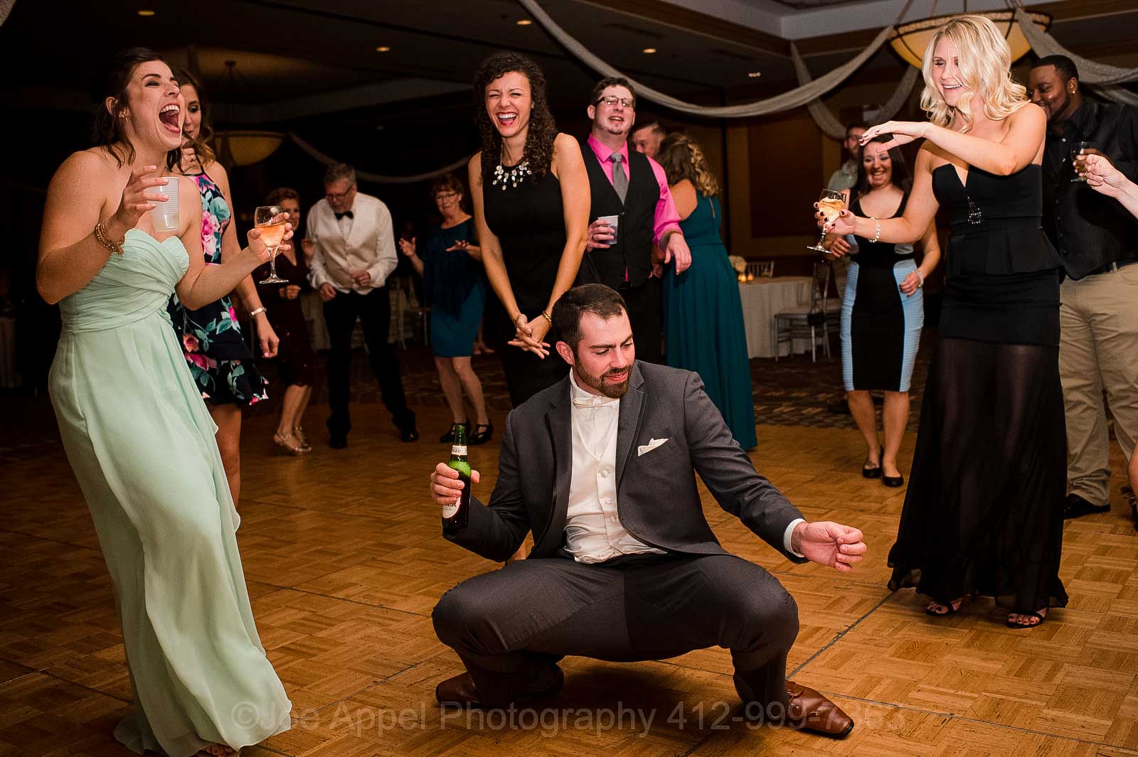 A groom holding a beer squats low and dances while guests laugh during his Embassy Suites Pittsburgh wedding.