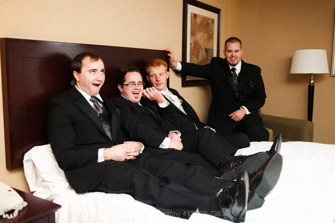 A groom and his groomsmen wearing black tuxedos clown around while sitting on a hotel room bed.