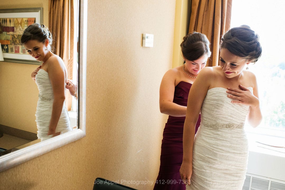 A bridesmaid zips up a bride's dress as she is reflected in the mirror in their hotel room.