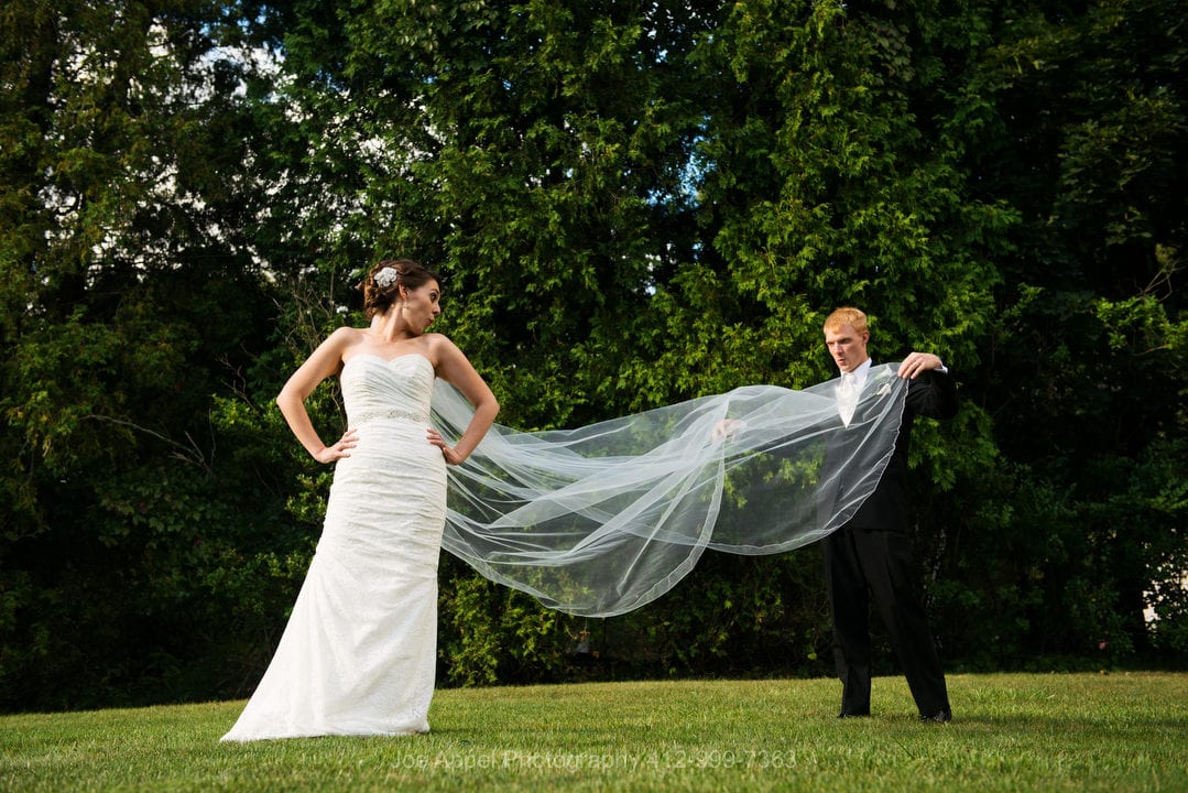 A bride and groom laugh as he holds out her long veil for a photo.