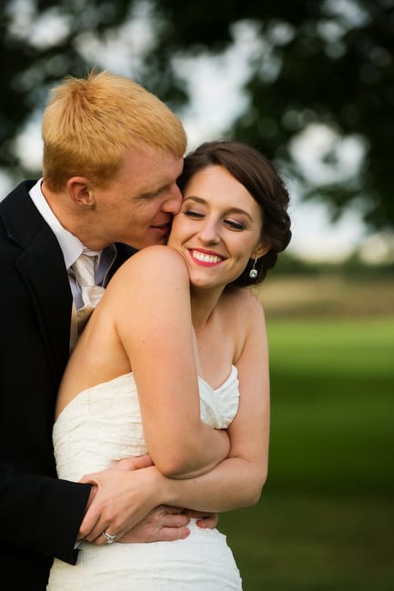 A groom holds on to his smiling bride from behind as he kisses her cheek.