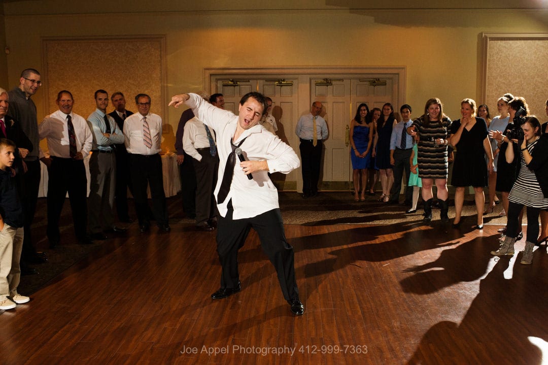 A man with a loosened tie and untucked shirt dances while surrounded by guests at a Montour Heights Country Club wedding.