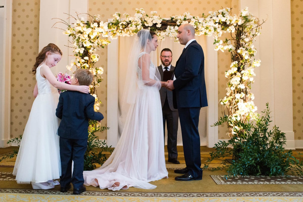 A bride and groom stand beneath a flowered arbor during their wedding ceremony indoors at Bedford Springs Resort