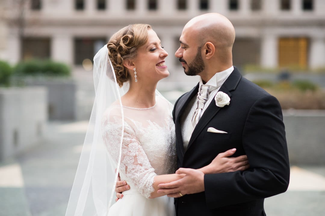 A bride in a lace sleeved gown stands with her bald headed groom wearing a tuxedo in front of the Omni William Penn in Pittsburgh.