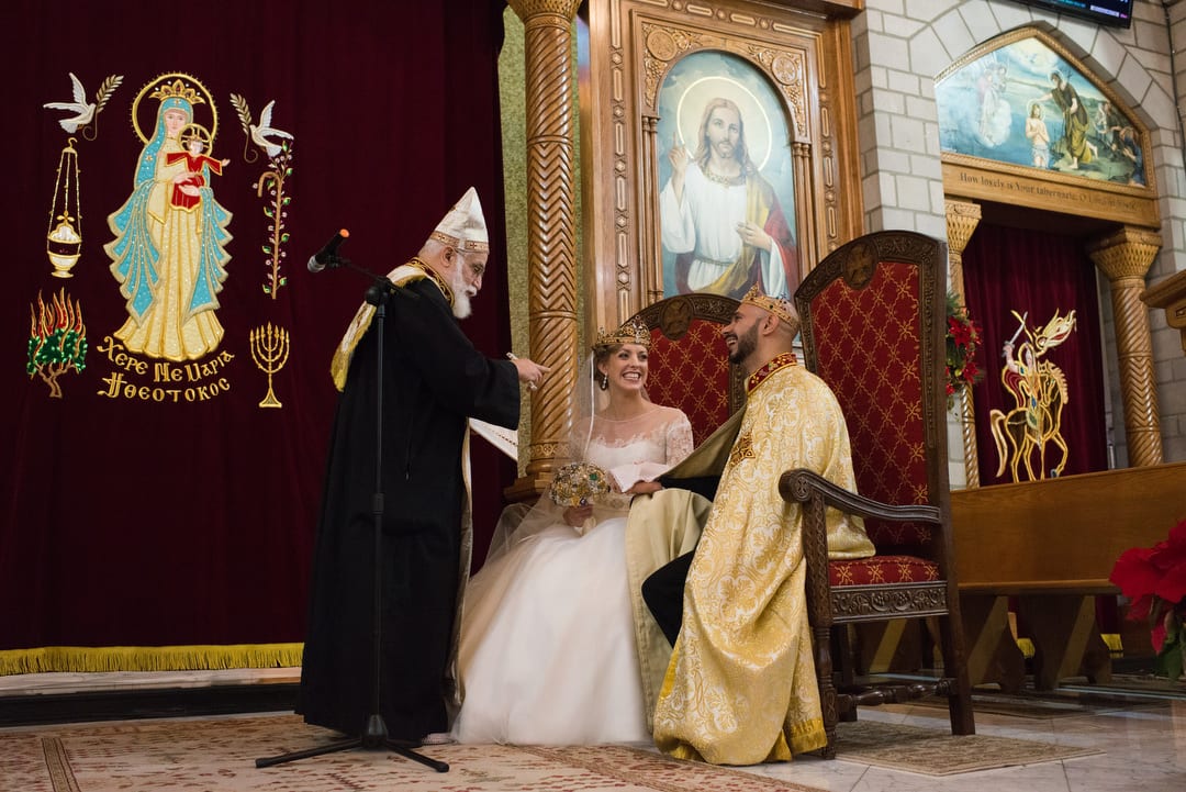 A priest blesses a bride and groom seated on large red upholstered chairs on the altar of St. Mary Coptic Orthodox church in Ambridge.