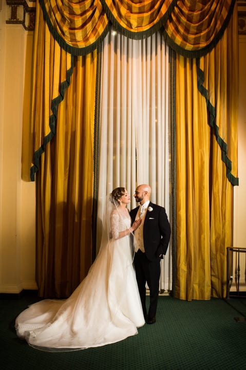 A bride and groom stand in front of the large gold-draped window in the lobby of the Omni William Penn hotel in Pittsburgh.
