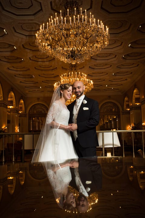 A bride and groom embrace as they and the chandeliers in the lobby of the Omni William Penn hotel are reflected in the glass table in front of them.