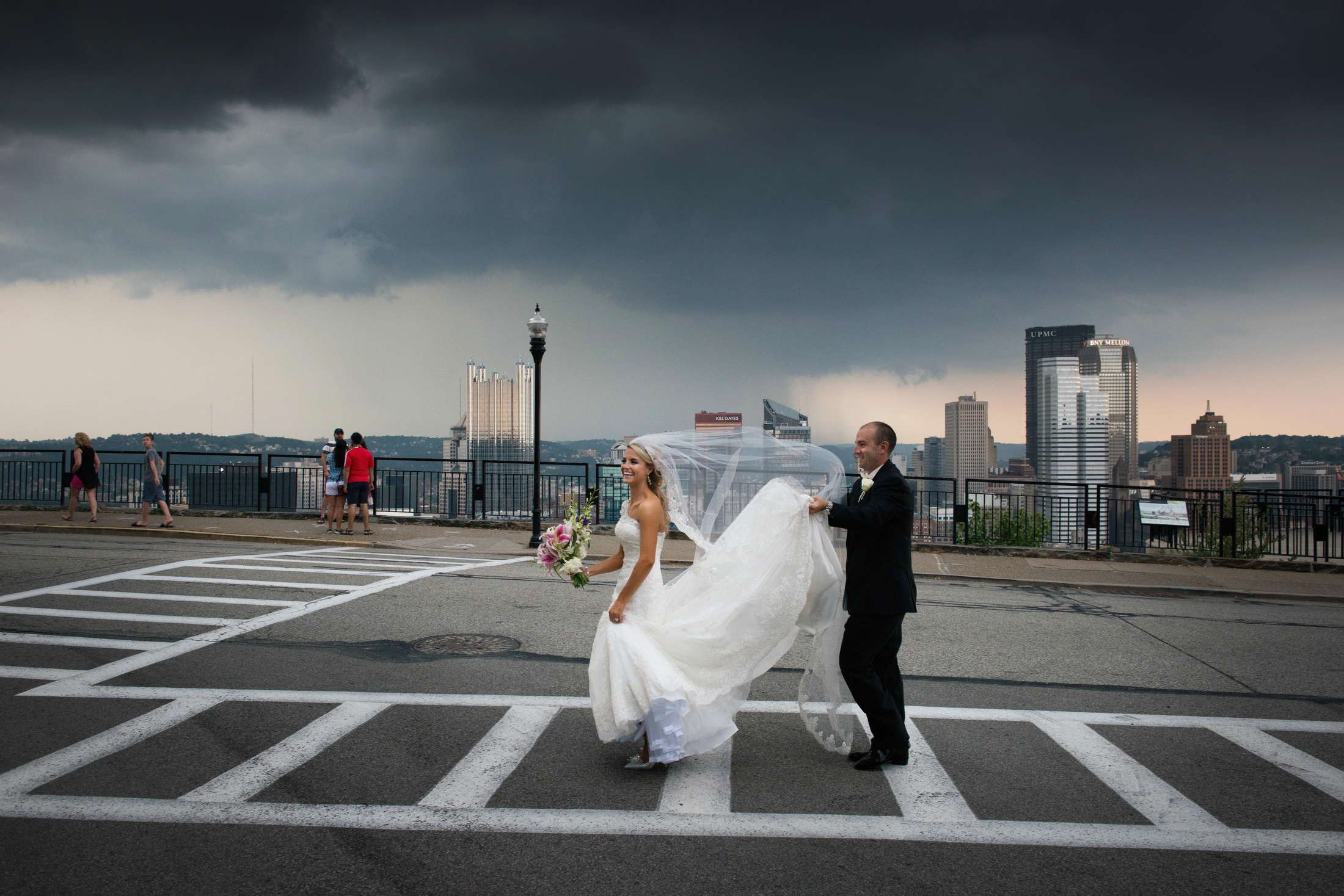 A bride and groom walk in a crosswalk on Grandview Avenue on Mt. Washington. The groom holds the bride's train while a storm approaches over downtown Pittsburgh.