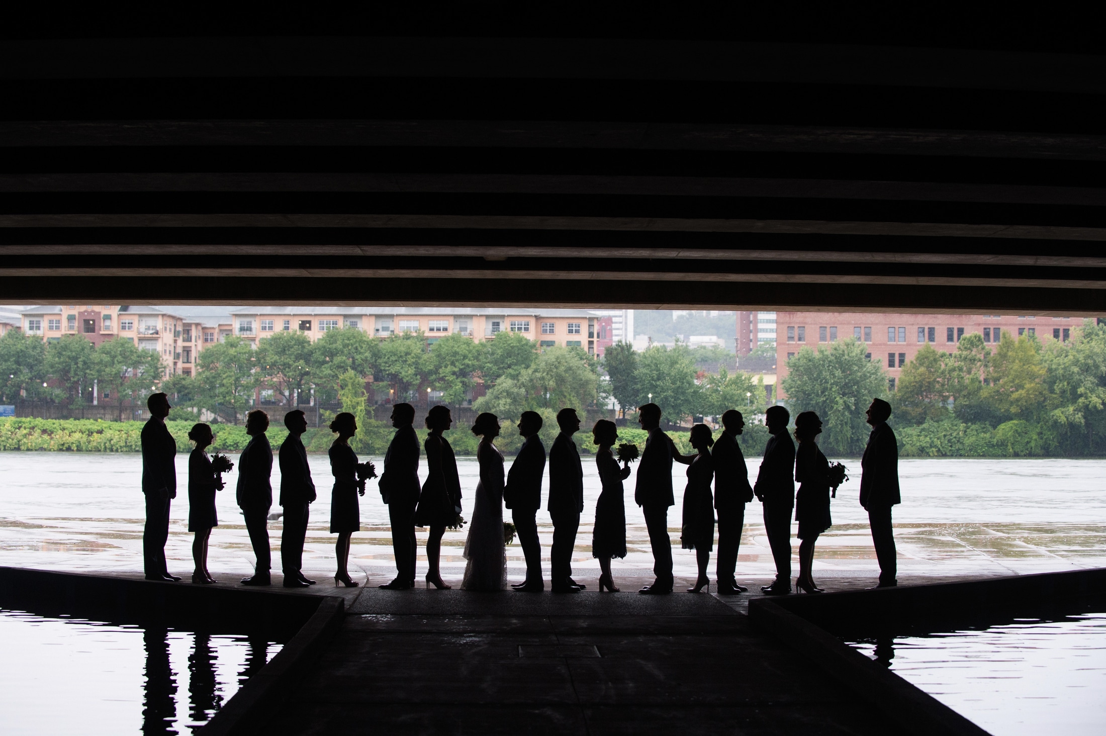 Seventeen members of a bridal party stand in silhouette beneath the convention center as they take wedding photos in the rain.