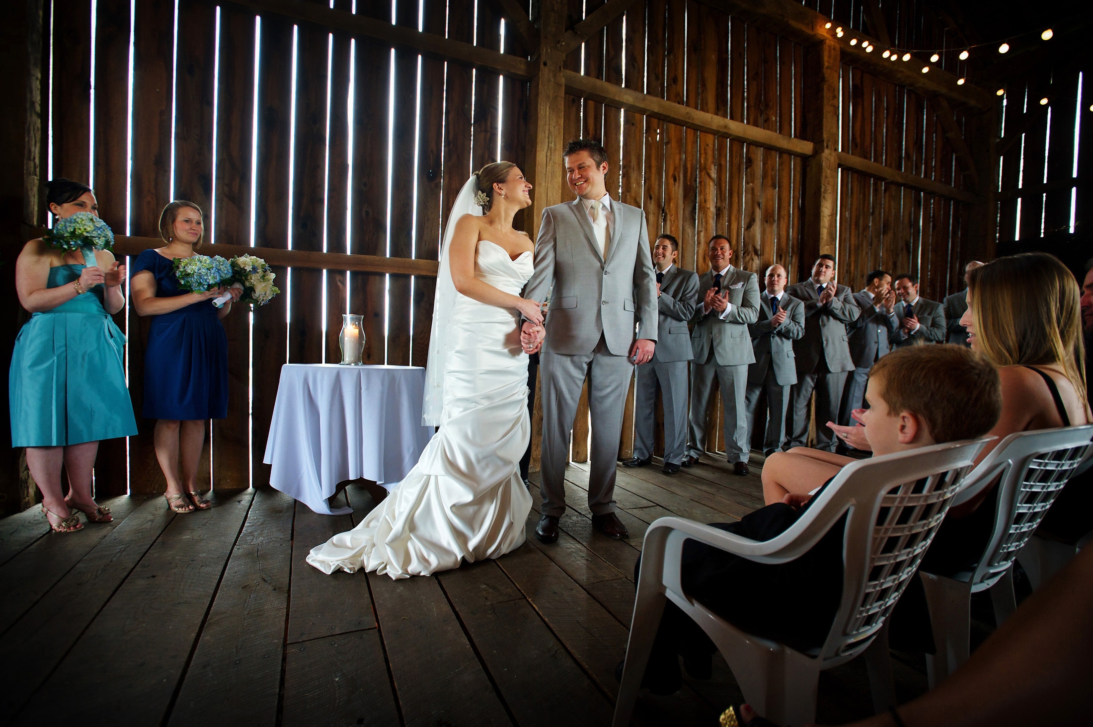 A bride and groom smile at each other at the end of their wedding ceremony inside of a barn on a rainy day.