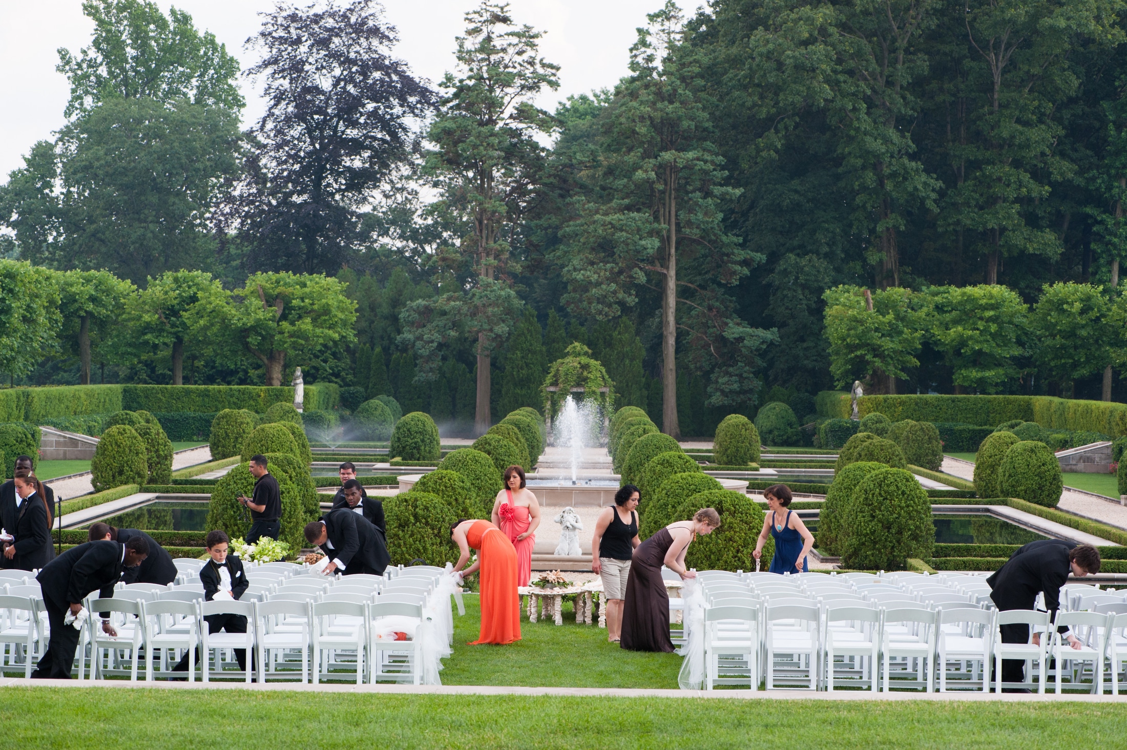 Wedding guests wearing bright gowns and tuxedos dry white folding chairs as they prepare for an outdoor ceremony after a rain storm.