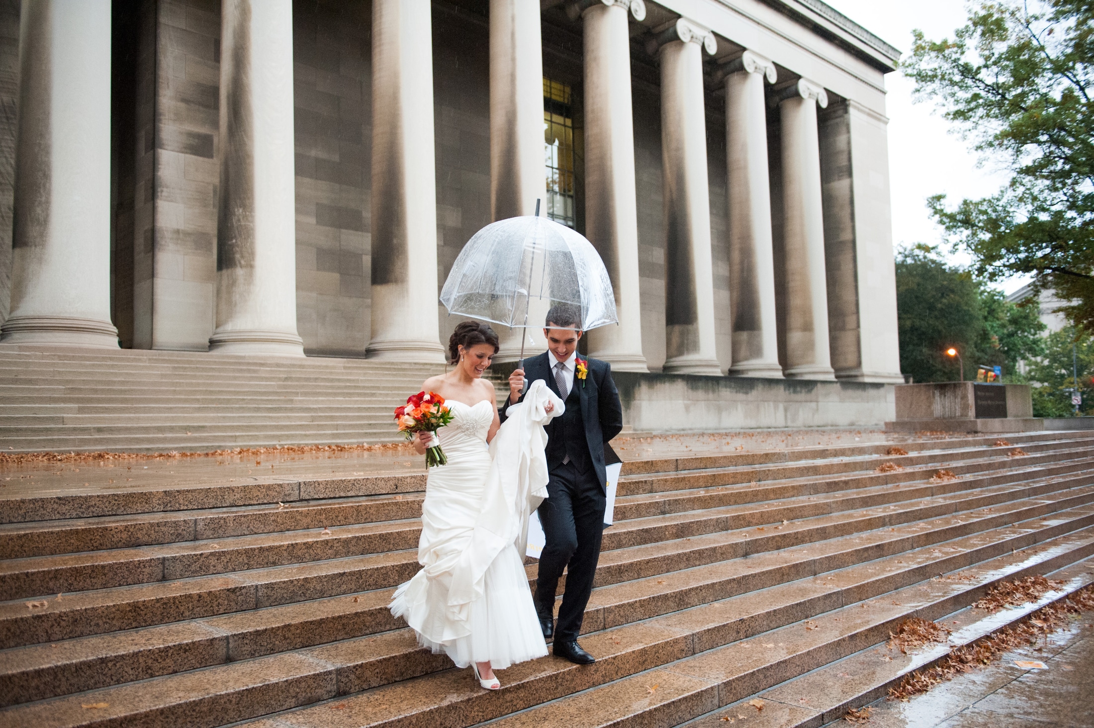 A bride and groom walk down the red granite steps in front of the Mellon Institute. She's holding a bright bouquet of flowers and he's holding a clear umbrella over them because it's raining. So we did wedding photos in the rain.