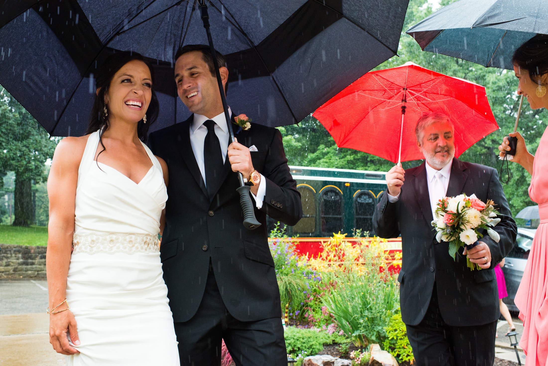 A bride and groom stand smiling together beneath a large golf umbrella as her father and sister stand beneath their own umbrellas during a rain storm.