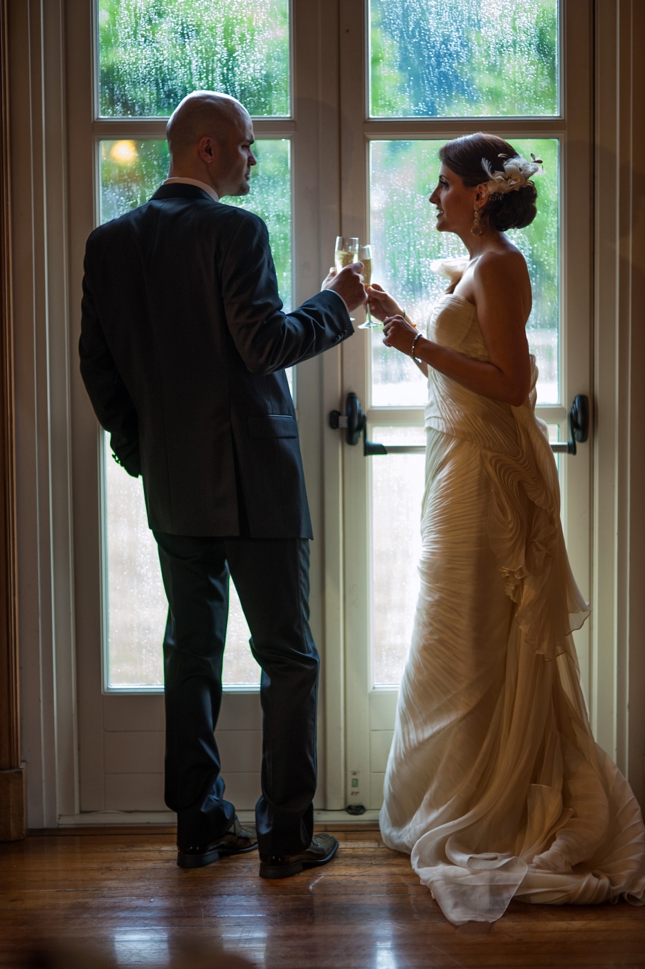 A bride and groom touch champagne flutes as they stand in front of a rain-soaked glass door.