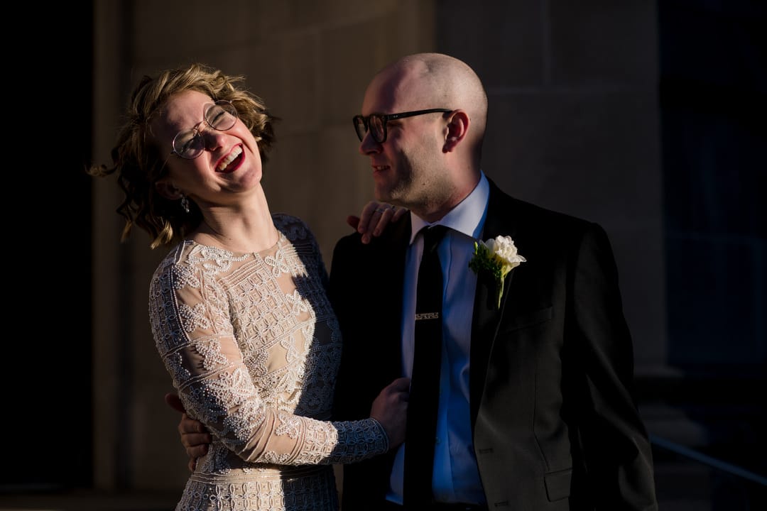 A blonde bride wearing glasses laughs as her groom looks at her and smiles in the late afternoon sun during their Ace Hotel Wedding.