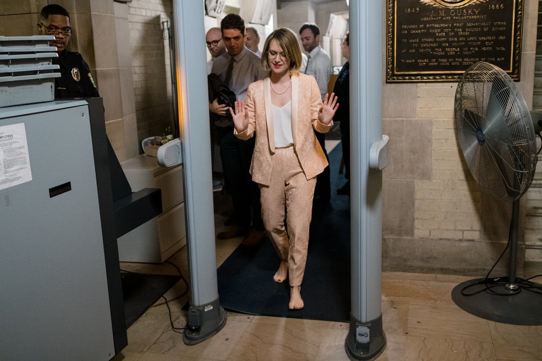 A woman in a pink pantsuit holds up her hands as she steps barefooted through a metal detector before her Allegheny County Courthouse wedding.