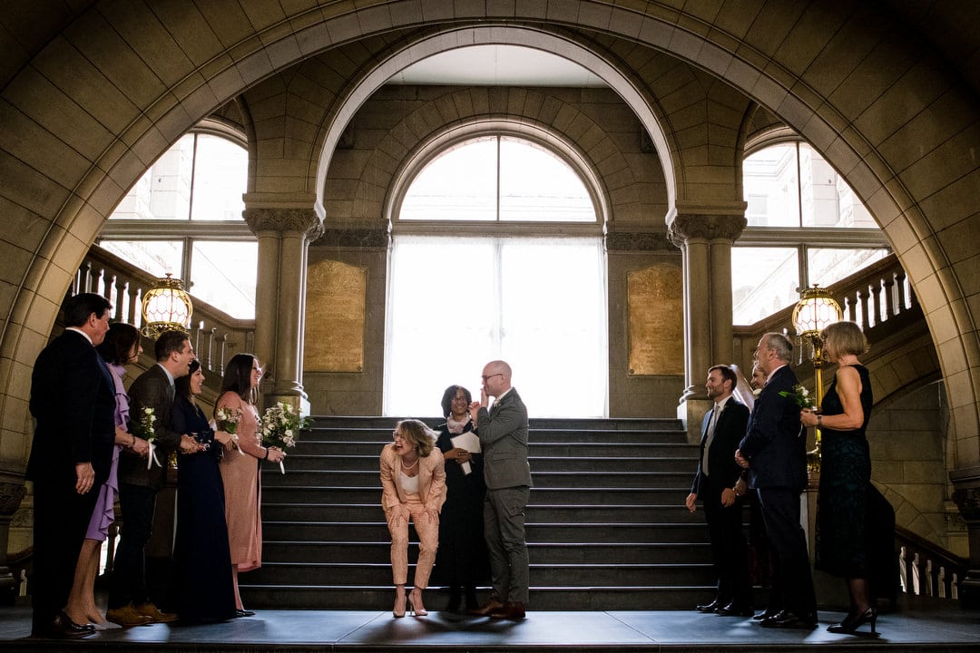 A bride in a pink pantsuit bends over and laughs as she turns to her family after kissing her groom beneath the arch in the stairway at the Allegheny County Courthouse.
