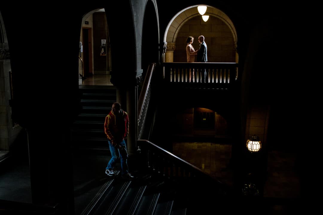 A couple faces each other as they are framed in an arch at the Allegheny County Courthouse. A man wearing a red jacket walks down the stairs in front of them.