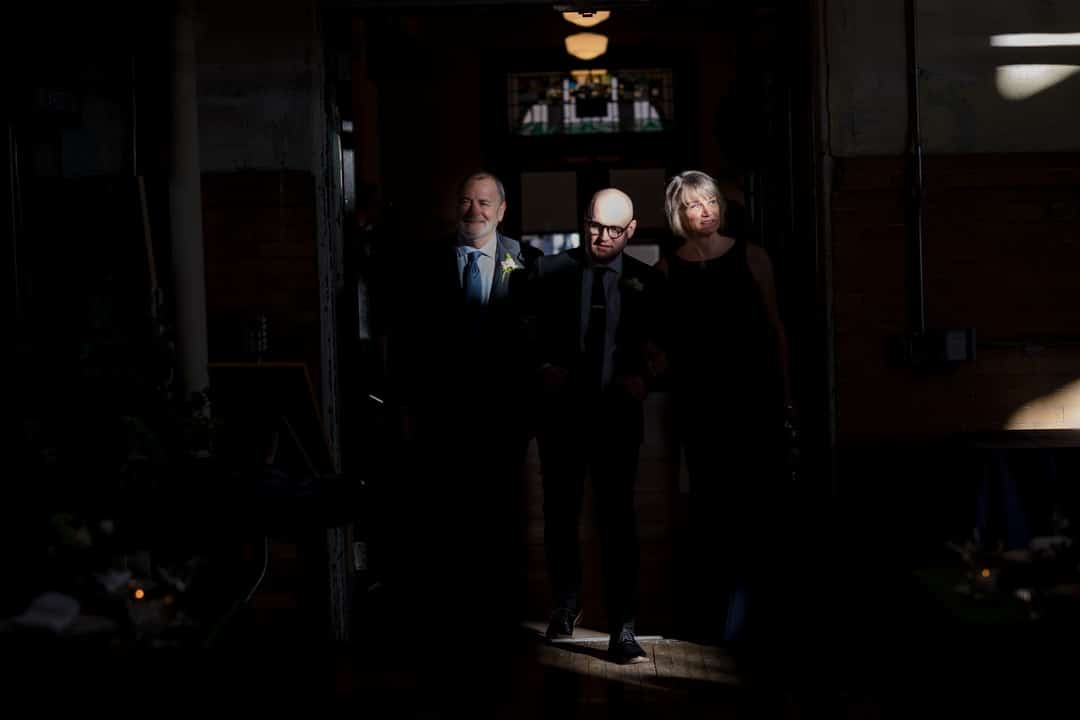 A groom is illuminated by a shaft of sunlight as he enters the gym with his parents during his Ace Hotel wedding.