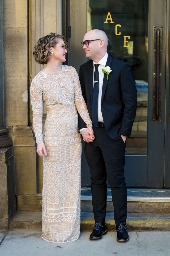 A bride and groom hold hands as they stand by the front door of the Ace Hotel Pittsburgh.
