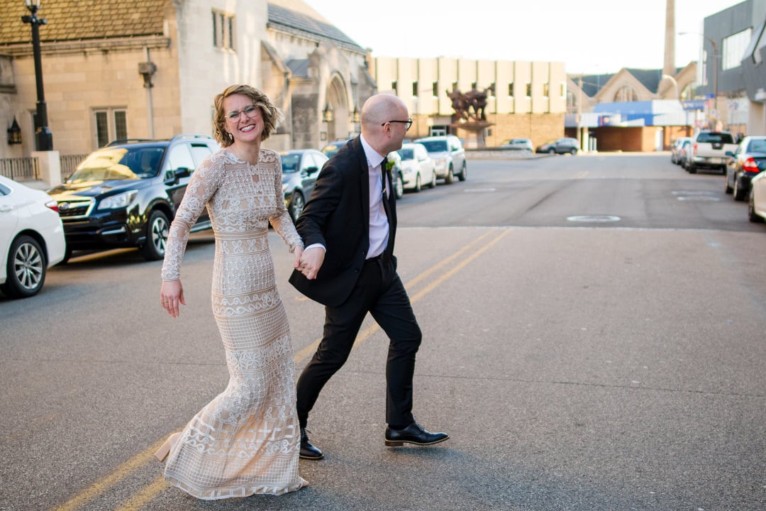 A grinning bride holds hands with her groom as they cross the street in front of the Ace Hotel Pittsburgh.