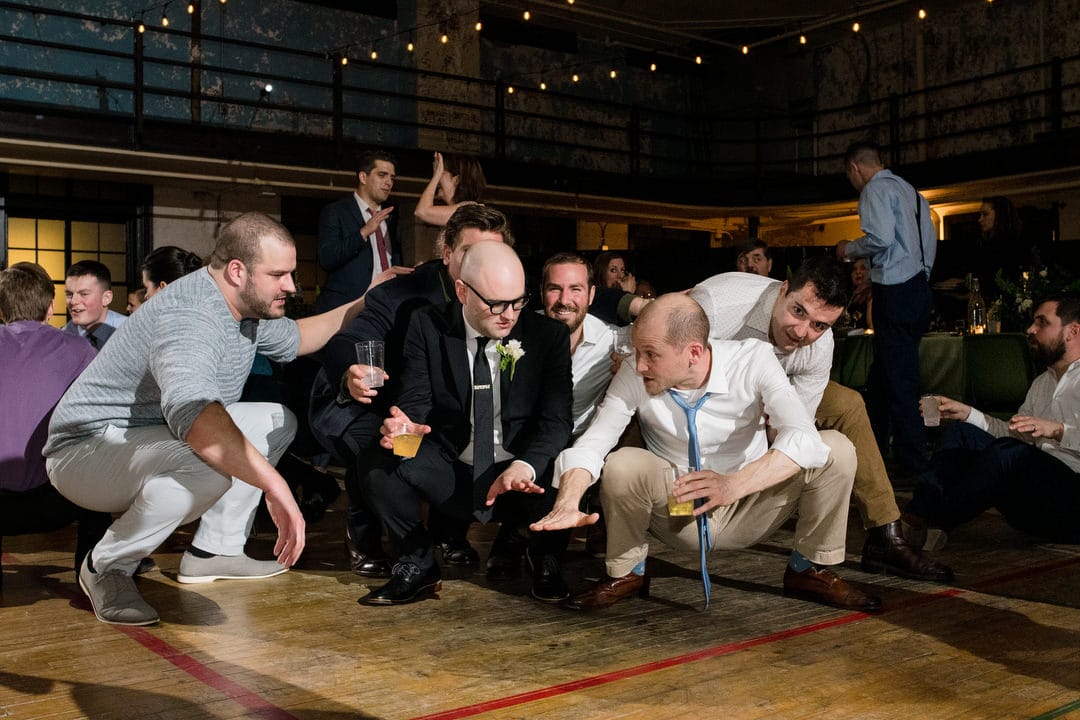 A groom wearing glasses, a dark suit, and with a shaved head squats with a group of friends as they dance during his wedding reception at the Ace Hotel Pittsburgh.