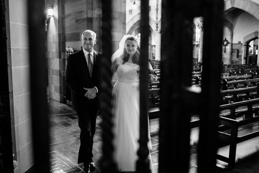 A bride and her father are seen through the gate to the Lady Chapel of Sacred Heart Church as they walk to her wedding ceremony.