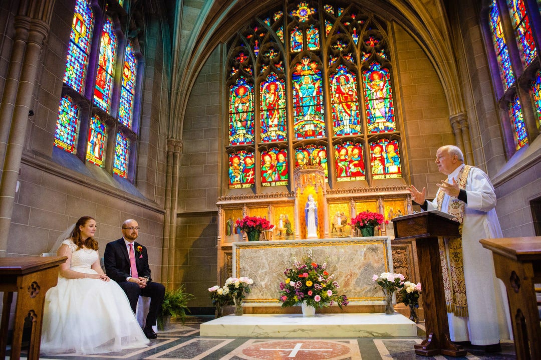 A bride and groom sit on the left side of the altar as a priest talks during their wedding ceremony inside of the Lady Chapel at Sacred Heart Church in Shadyside.