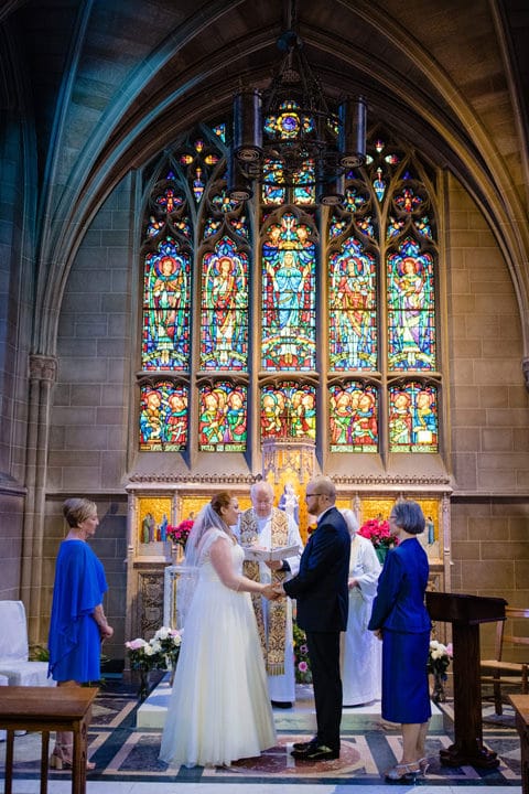 A bride and groom hold hands during their wedding ceremony as their mothers stand by their sides at Sacred Heart Church in Shadyside.
