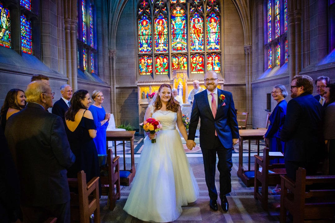 A bride and groom smile as they walk out of their wedding ceremony in the Lady Chapel at Sacred Heart while their guests applaud.
