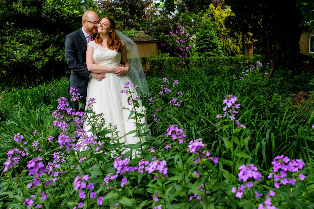 A groom embraces his red haired bride from behind as they stand in a patch of purple flowers.