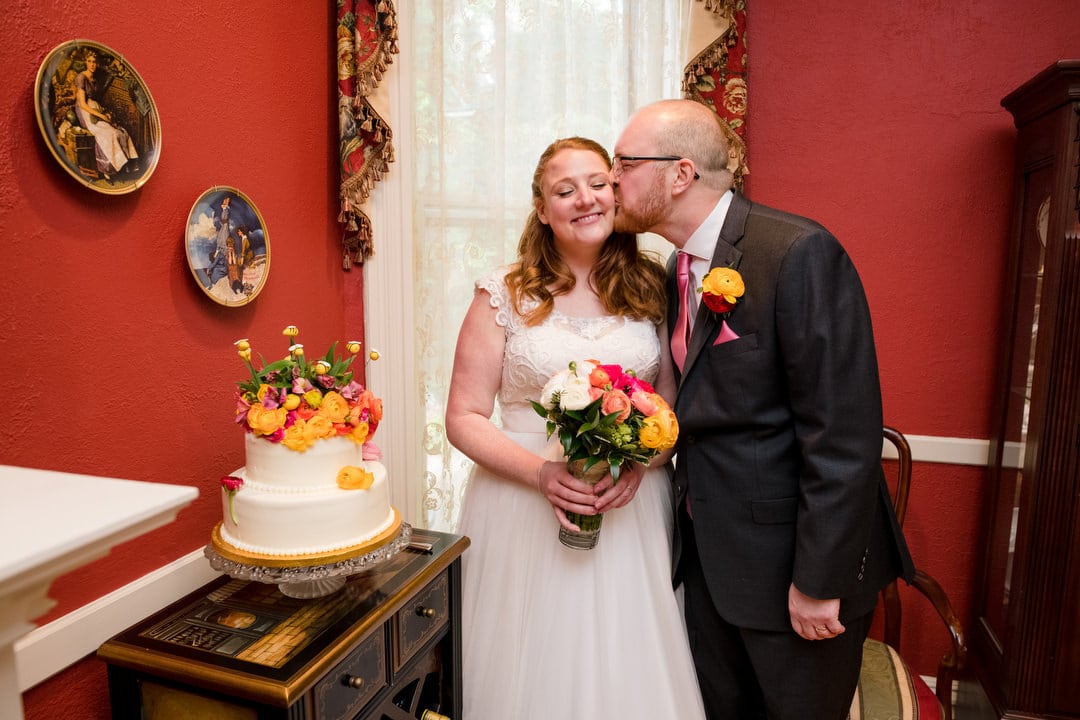 A groom kisses his bride on the cheek as they stand next to their wedding cake in a room with red walls.