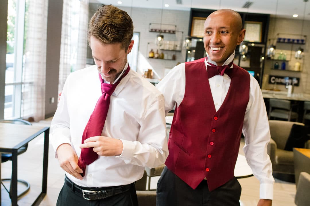 A groom wearing a white shirt and red vest helps a groomsman as he ties his red tie.