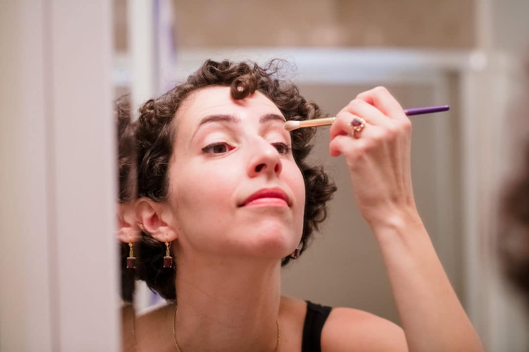 A woman with brown curly hair applies eye makeup with a brush in a mirror.
