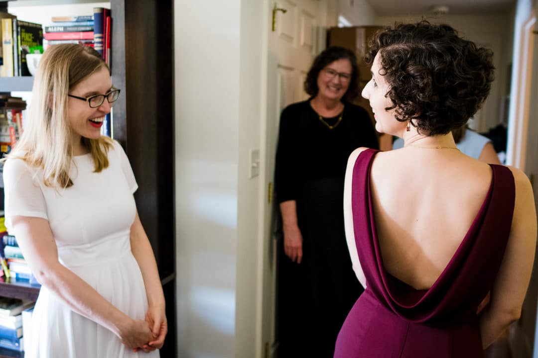 A bride wearing a dark crimson dress smiles with her bridesmaids as she shows them the back of the dress.