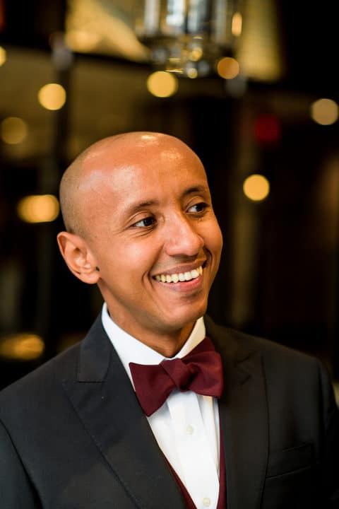 A groom with a shaved head smiles as he looks to the right. He is wearing a dark suit and a crimson tie.