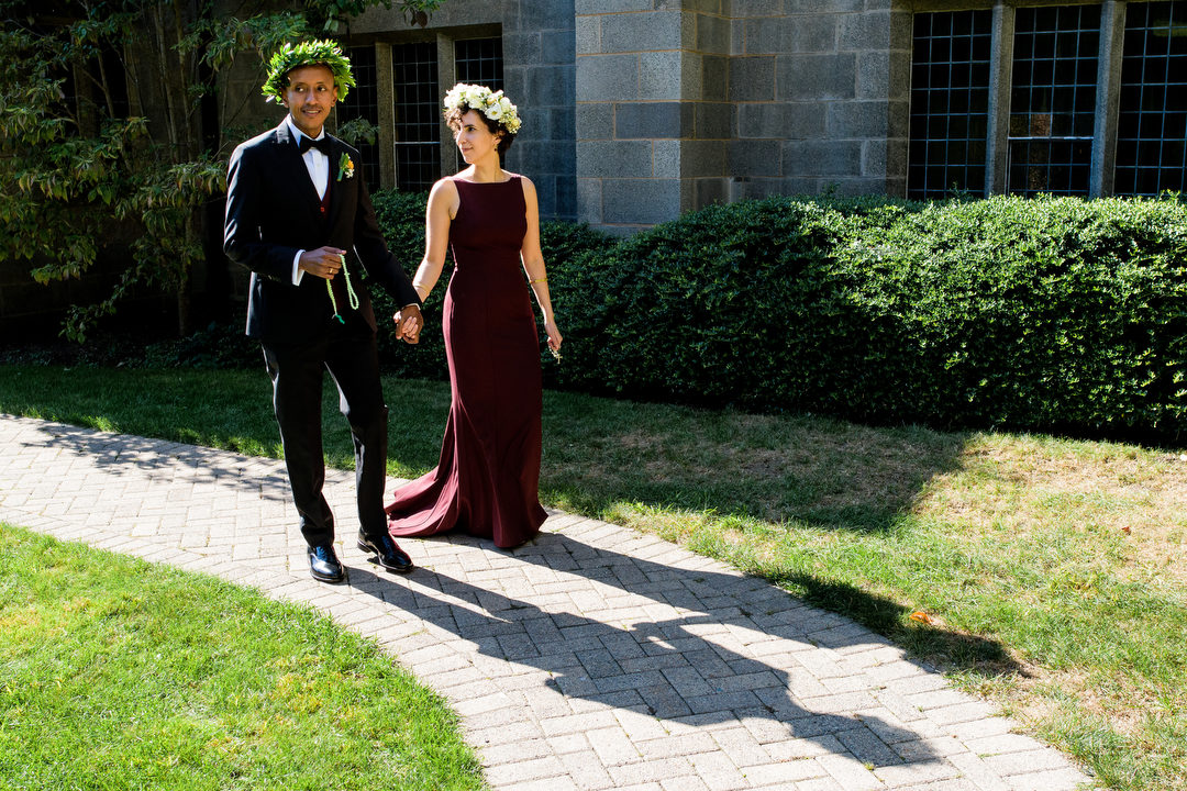 A bride and groom hold hands as they walk on a path while wearing flower crowns.
