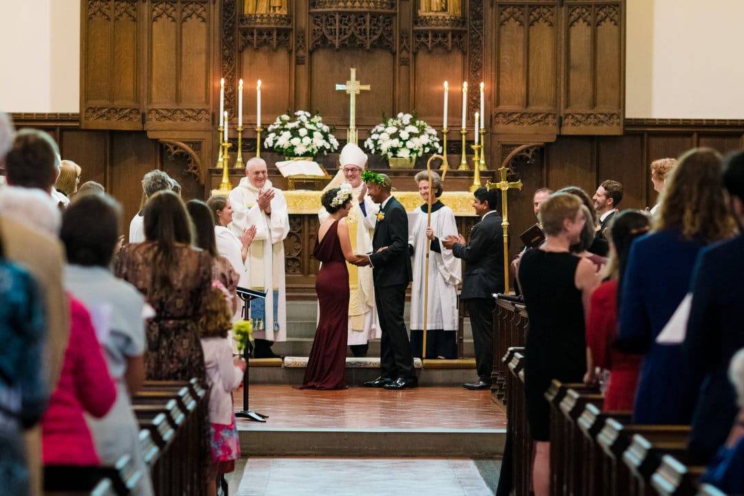 A bride and groom hold hands as they are blessed by the Episcopal Bishop at the Church of the Redeemer in Chestnut Hill MA
