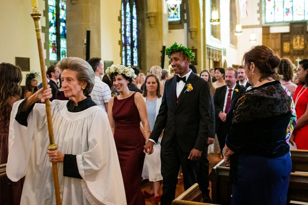 Led by a woman wearing vestments holding a cross, a couple recesses down the aisle at the end of their wedding ceremony.